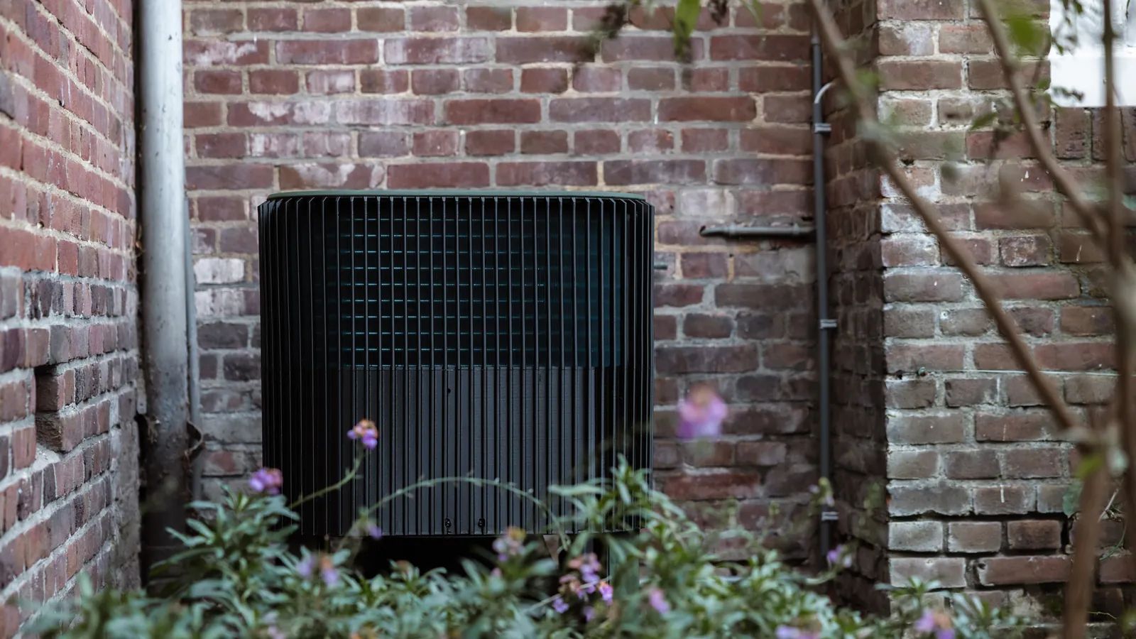 De buitenunit van de Whspr-warmtepomp staat tegen de bakstenen achtergevel van een woonhuis, omringd door tuinplanten.